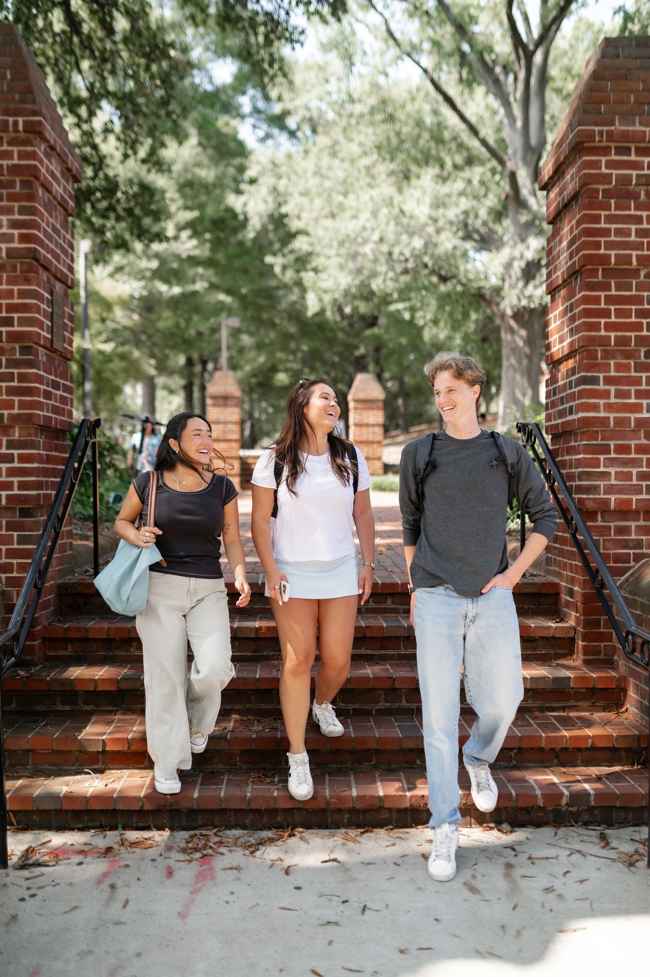 Three students are walking down brick steps outdoors, smiling and talking. They are carrying bags and appear happy, surrounded by trees and brick pillars on a sunny day.
