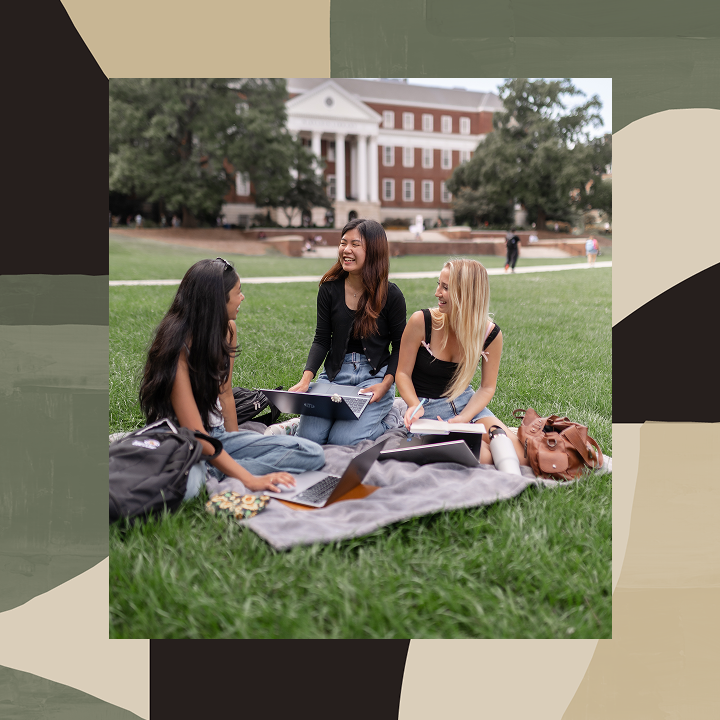 Three young women sit on the grass with notebooks and laptops, smiling and talking on a college campus with a red-brick academic building and trees in the background.