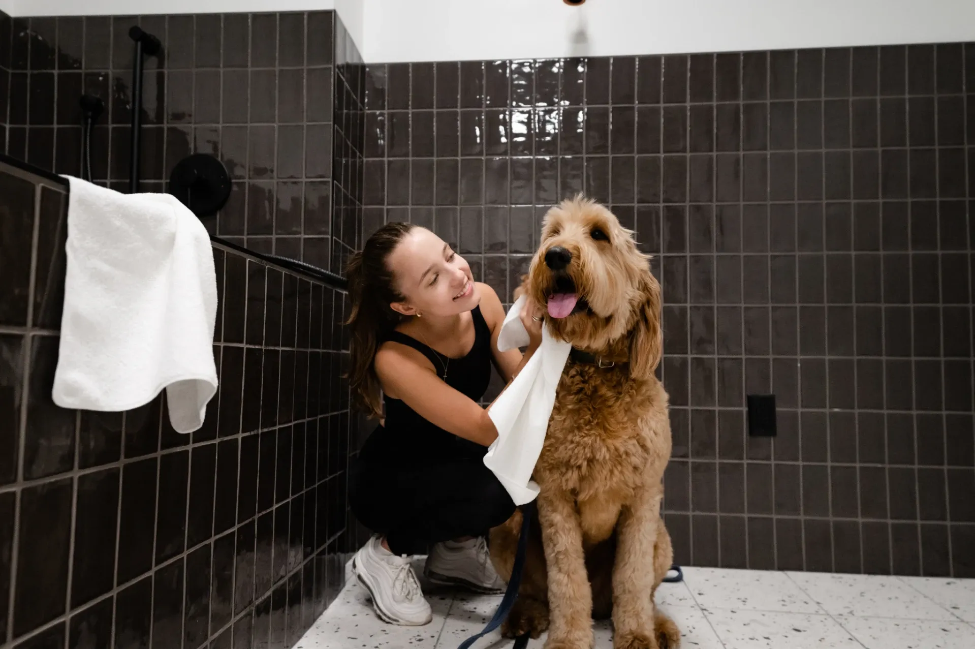 A woman kneels beside a large, fluffy dog in a tiled bathroom at University of Maryland housing, smiling as she dries the happy pup with a white towel. The dog's tongue hangs out cheerfully.
