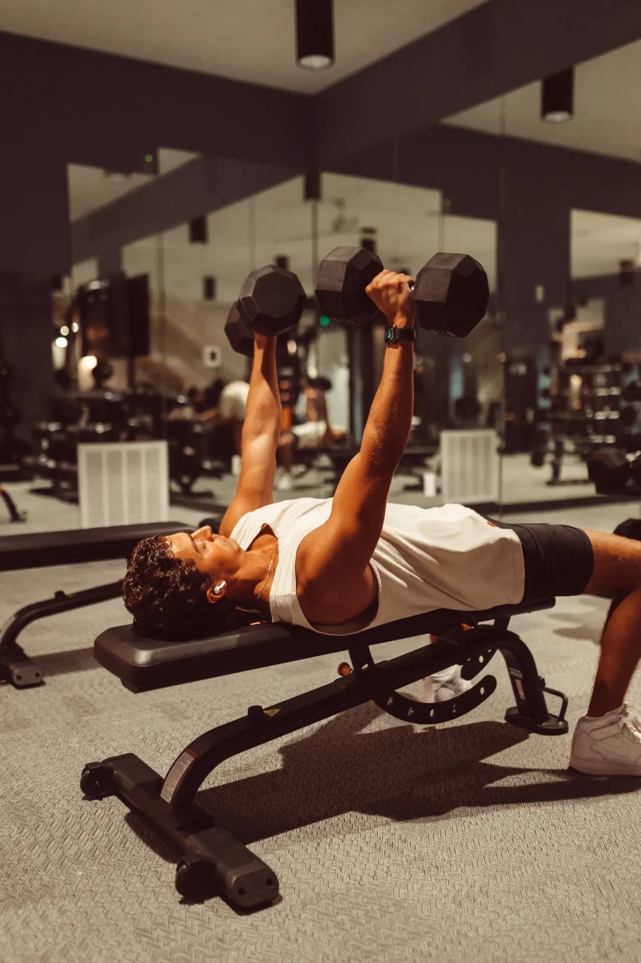 A person in a white tank top and shorts is lying on a bench in a University of Maryland housing gym, lifting dumbbells above their chest. Gym equipment and mirrors are visible in the background.