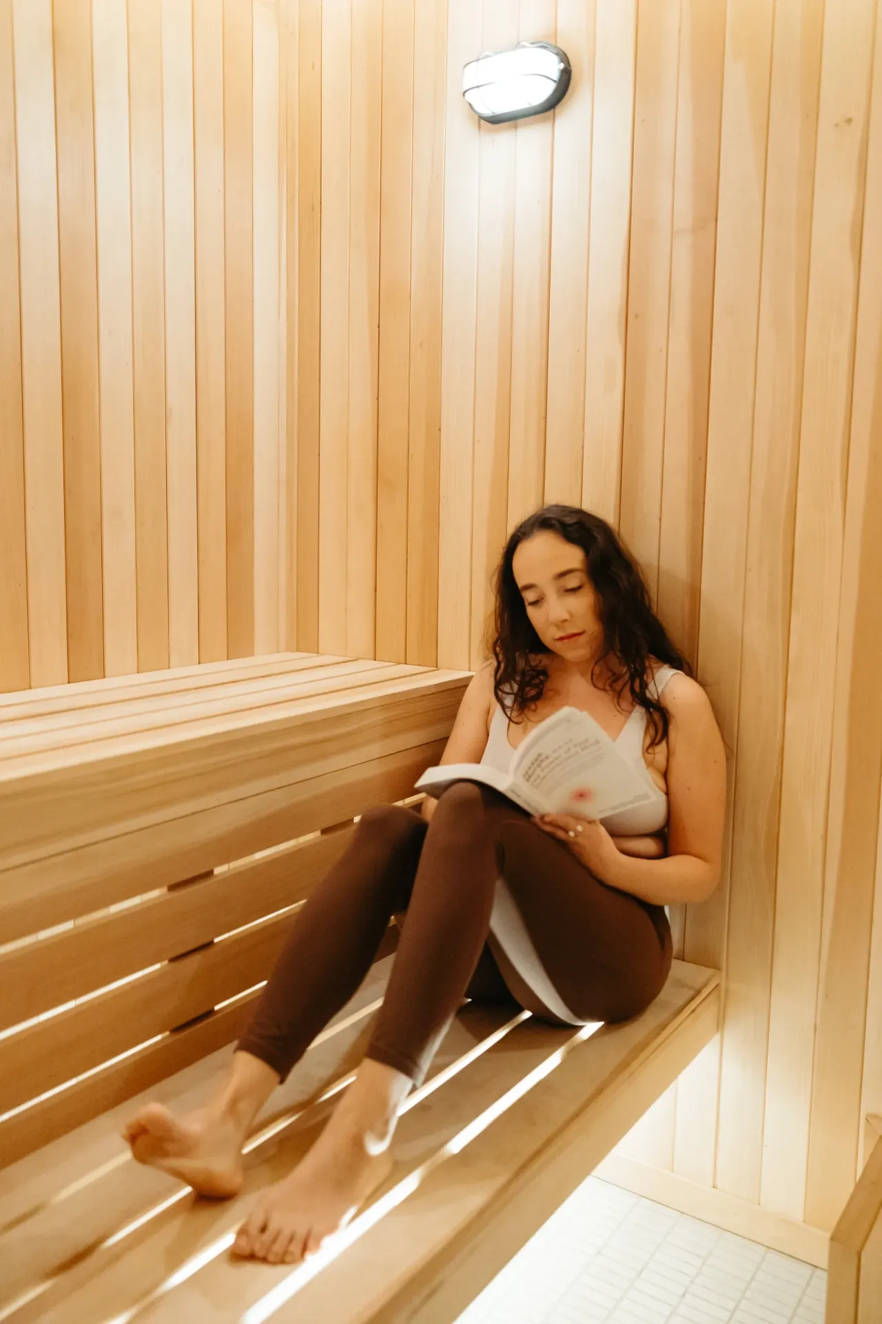 A woman in a swimsuit and leggings sits on a wooden sauna bench, reading a book—perhaps studying University of Maryland housing options. Warm light illuminates the wooden walls and benches around her.