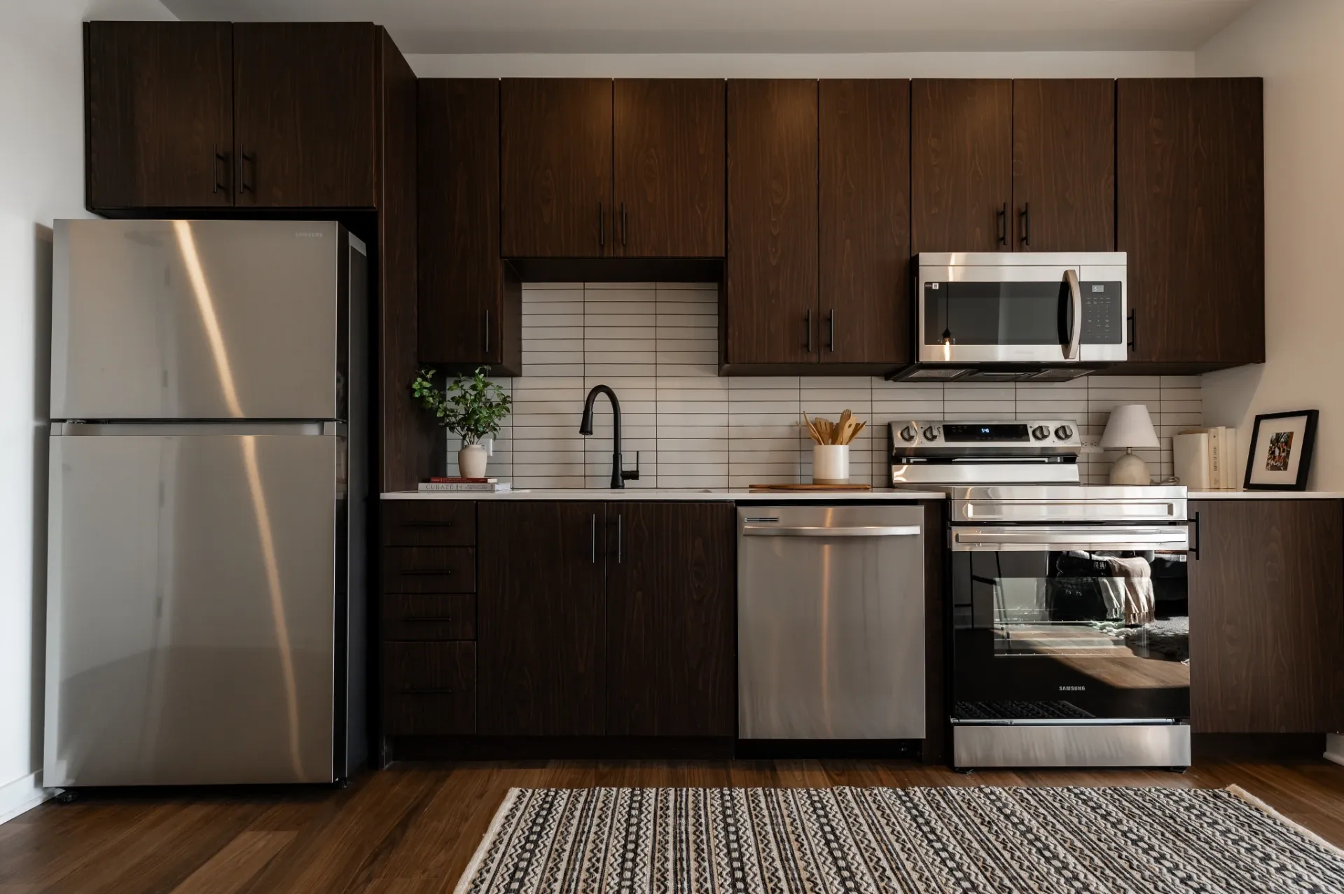 Modern kitchen in University of Maryland housing featuring dark wood cabinets, stainless steel appliances—fridge, dishwasher, oven, microwave—white tile backsplash, a plant and a striped rug on a wooden floor.