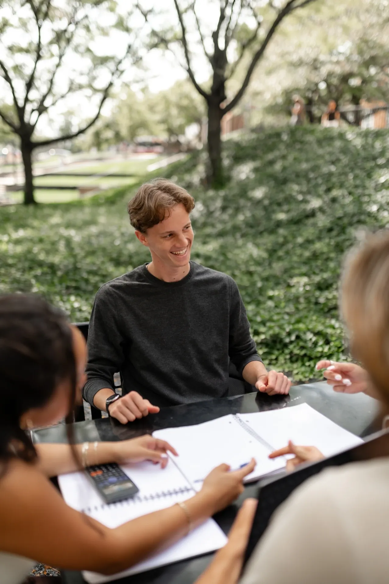 Three students sitting at an outdoor table with open notebooks and a calculator, discussing University of Maryland housing. One person in a dark shirt is smiling as two others gesture toward the papers. Green foliage forms a calm backdrop.
