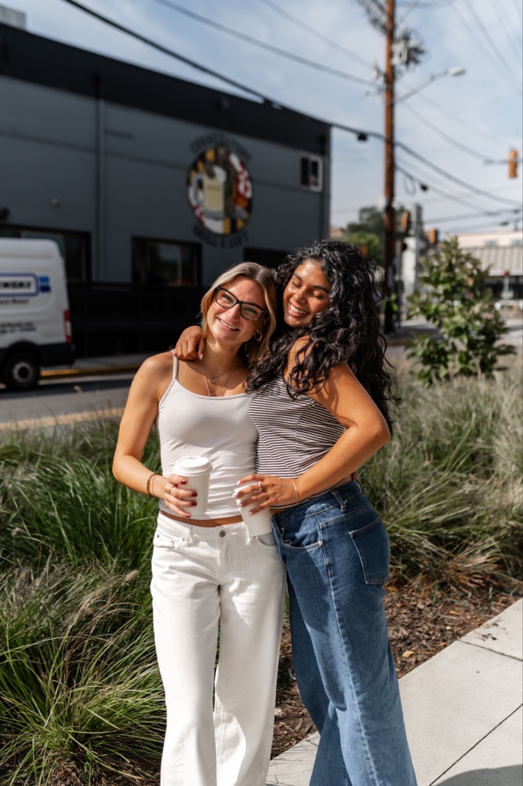Two women stand outside on a sunny day, smiling and hugging each other—almost like friends who've just finished finding a roommate. One wears a white tank top and pants; the other wears a striped top and blue jeans, with urban buildings and greenery behind them.