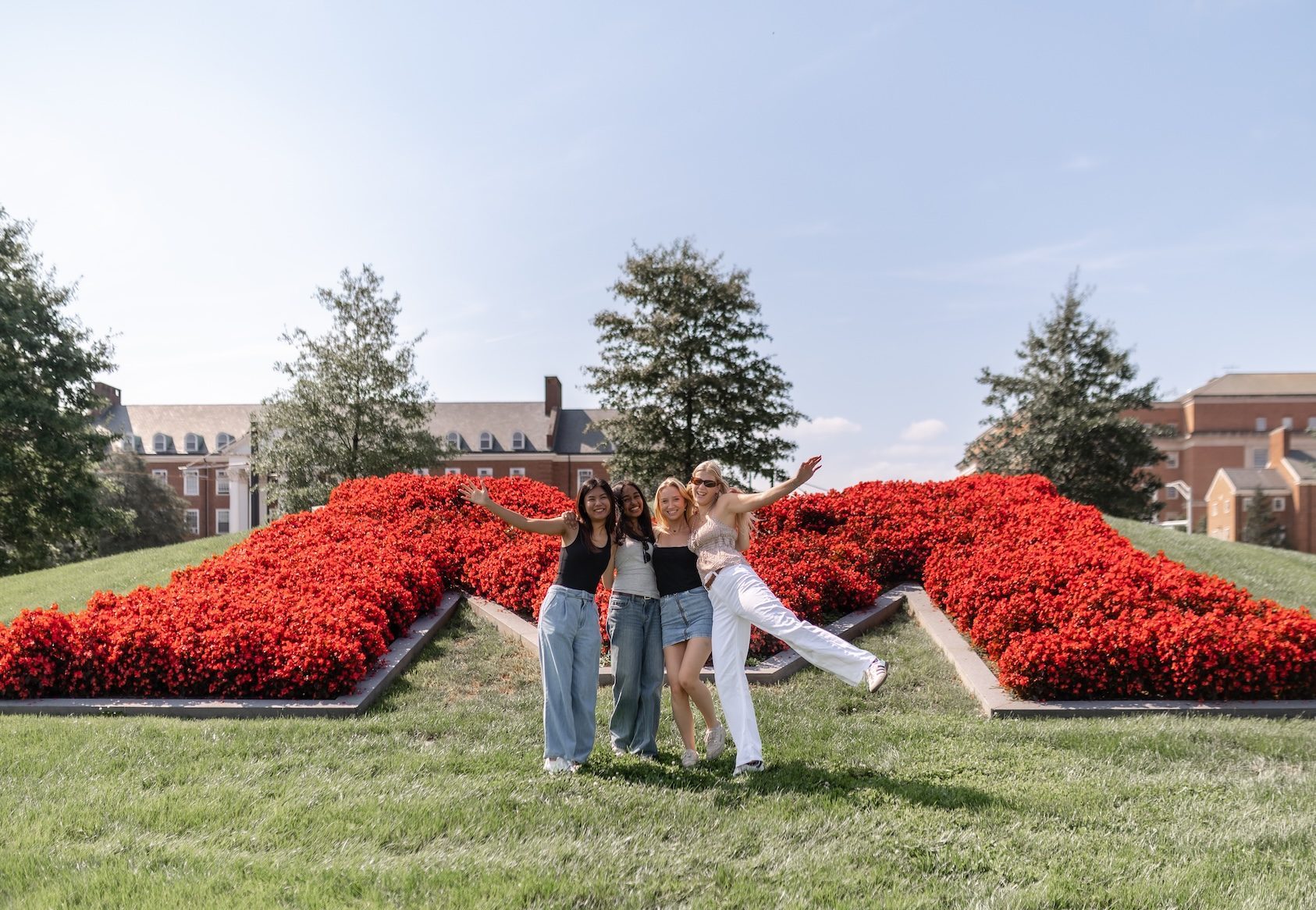 Three people pose happily in front of a large red M made of flowers on a grassy hill in College Park, with campus buildings and trees in the background—a perfect spot for those seeking student apartment jobs nearby.