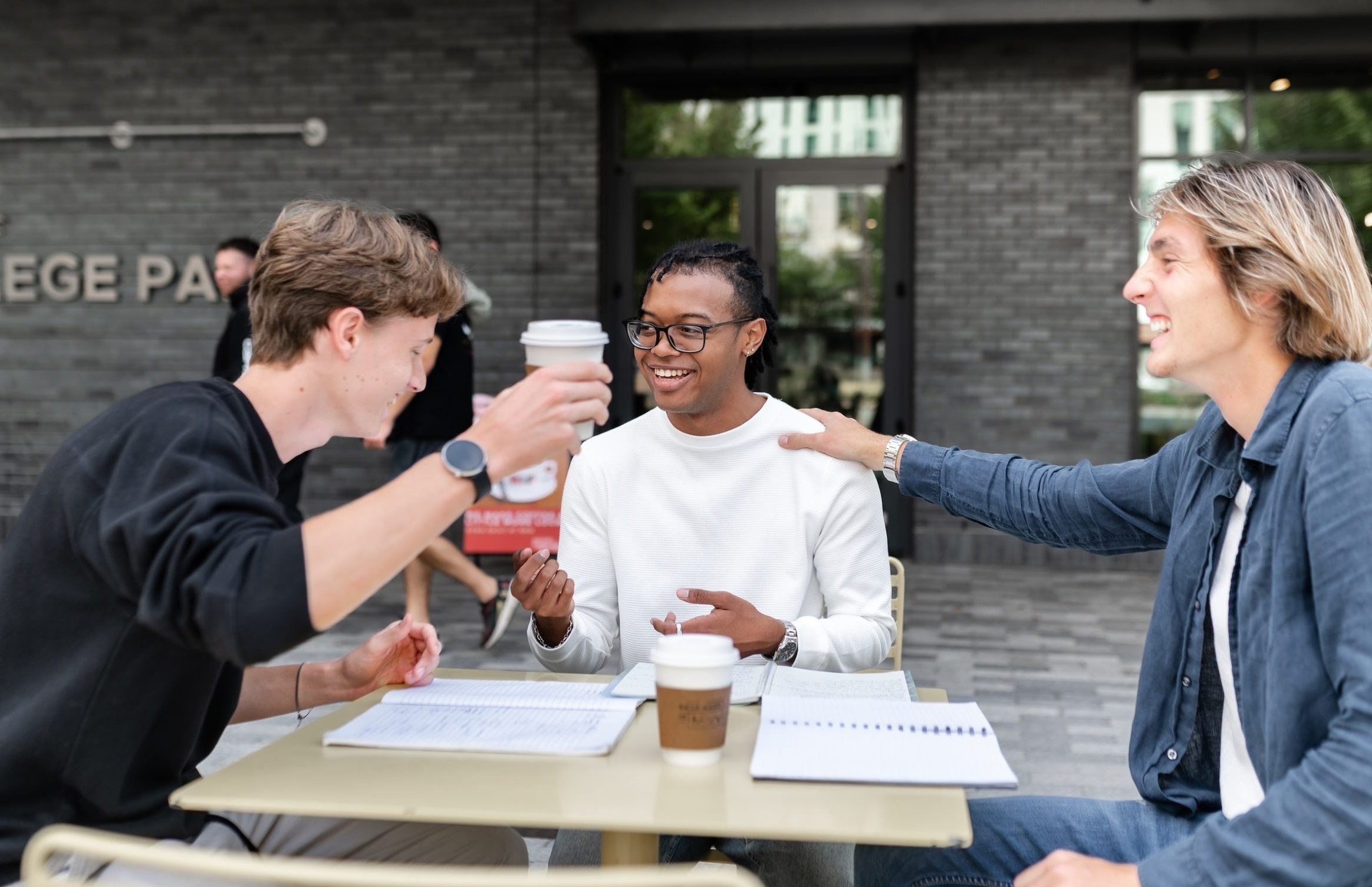 Three people sit at an outdoor table in College Park, smiling and talking. Two have coffee cups, and one raises a cup in a cheerful gesture. Notebooks and papers suggest they're discussing student apartment jobs or study plans.