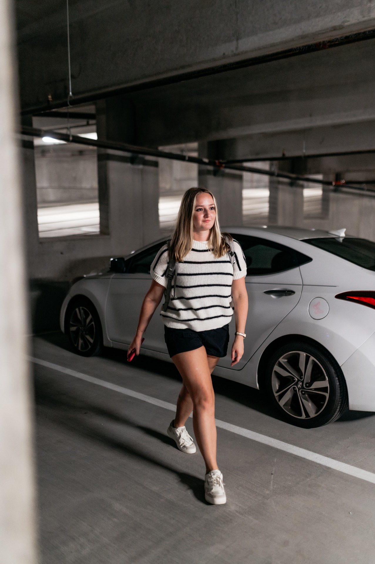 A woman in a striped shirt, black shorts, and white sneakers walks confidently in a parking garage next to a white car, holding a phone in her hand.