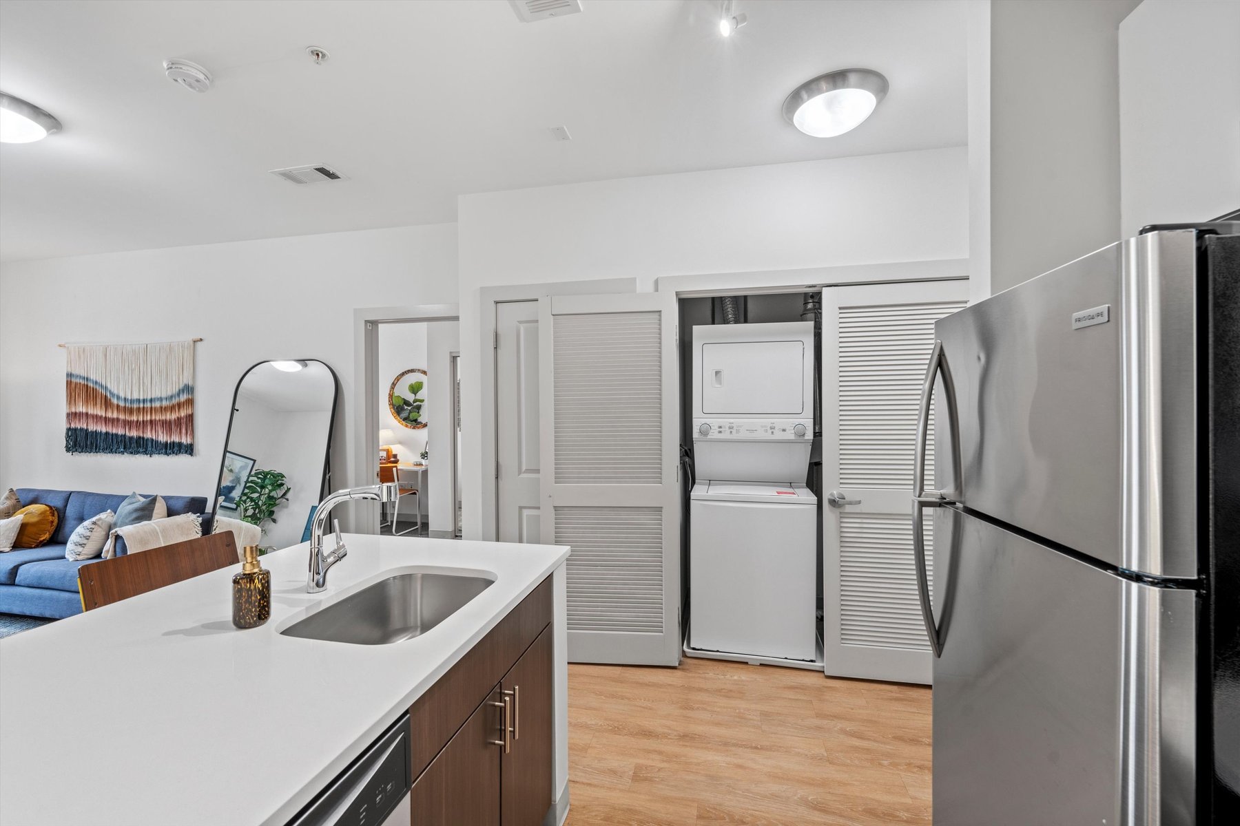Modern apartment kitchen with stainless steel appliances and a sink island at Rambler College Park South; in the background, an open closet reveals a stacked washer and dryer, plus a living area with a blue sofa and wall art.