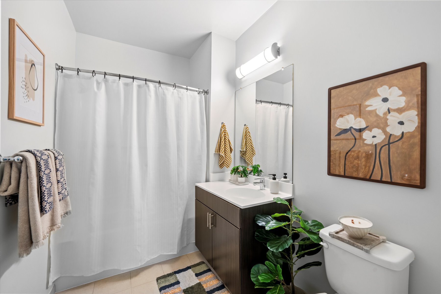 Modern bathroom at Rambler College Park South features a white shower curtain, dark vanity with sink, large mirror, towel rack, toilet, potted plant, and flower-themed wall art. Towels and a striped rug add color to the neutral space.