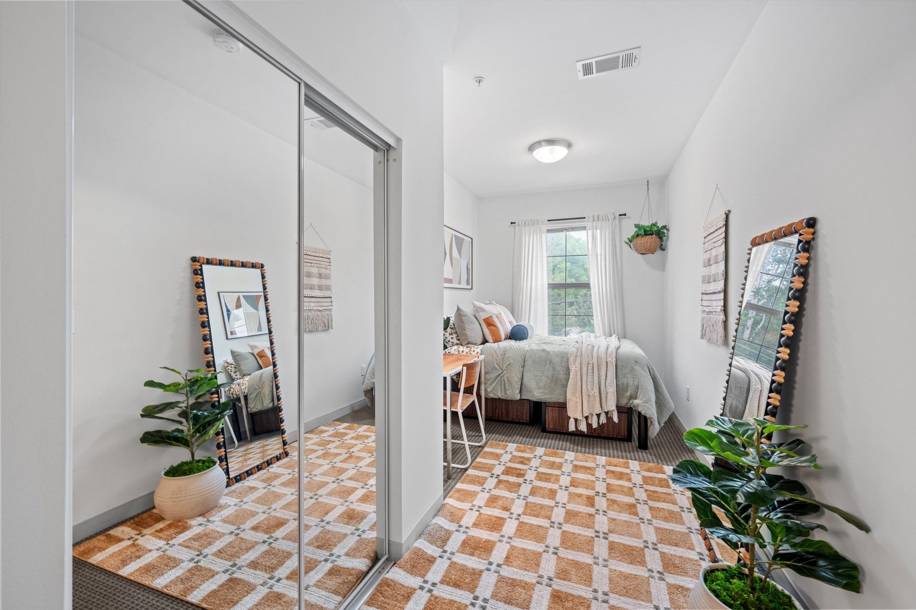 A bright, modern bedroom at Rambler College Park South features a bed, desk, chair, potted plants, large mirror, and a window with sheer curtains. Earthy tones and a checkered rug complete the look, with mirrored closet doors on the left.