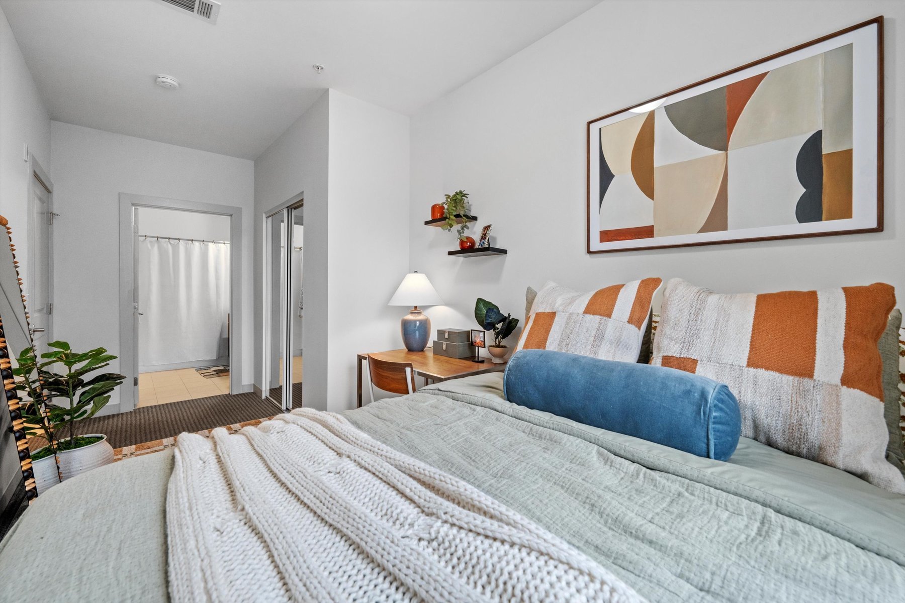 A cozy bedroom at Rambler College Park South features a neatly made bed with striped pillows and a blue bolster. A desk with a lamp and plants sits against the wall, modern art hangs above the bed, and a bathroom is visible through an open doorway.