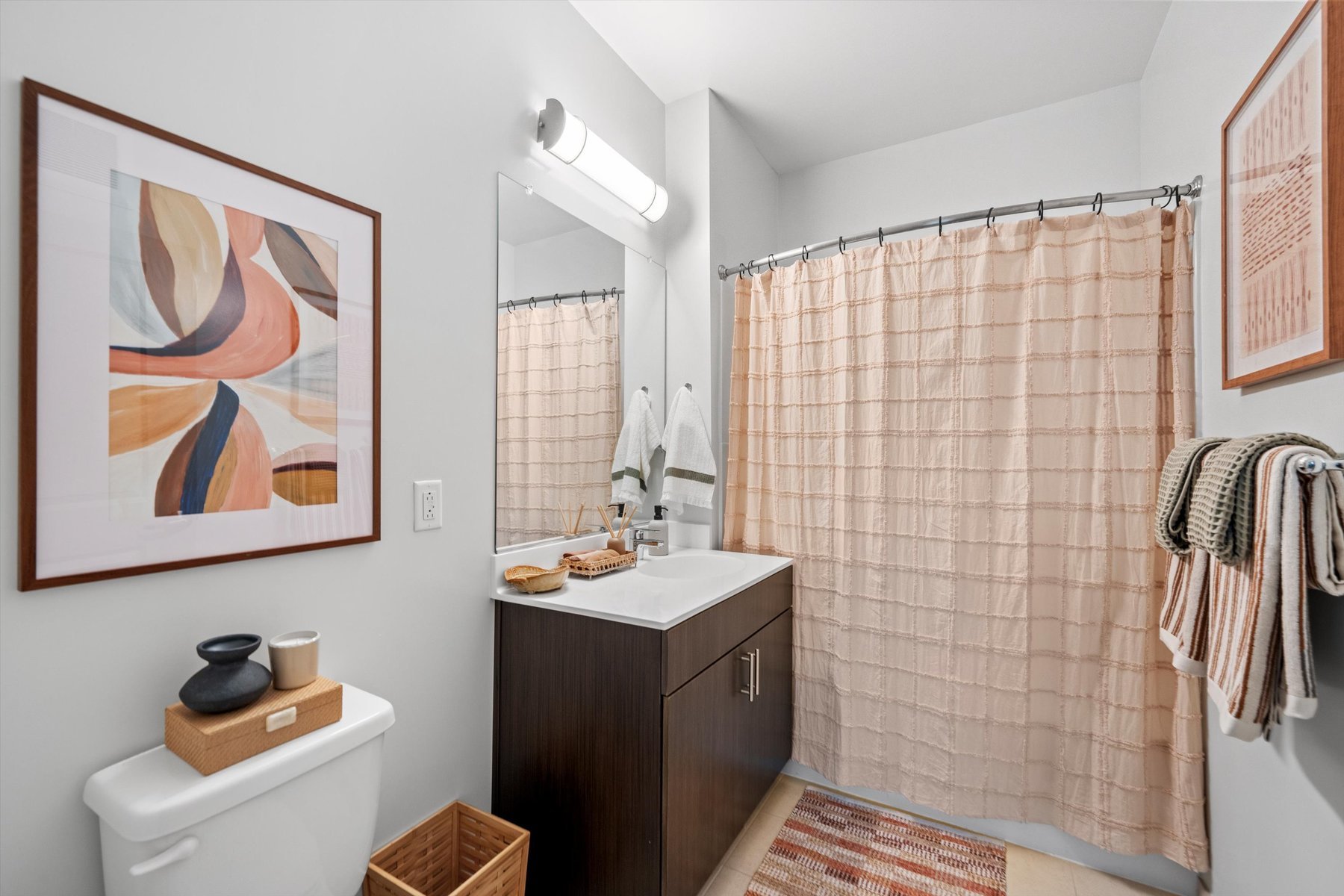 Modern bathroom at Rambler College Park South with a dark wood vanity, white sink, large mirror, and overhead light. Abstract art hangs above the toilet. The shower has a peach curtain, and towels hang on wall hooks in this warm, neutral space.