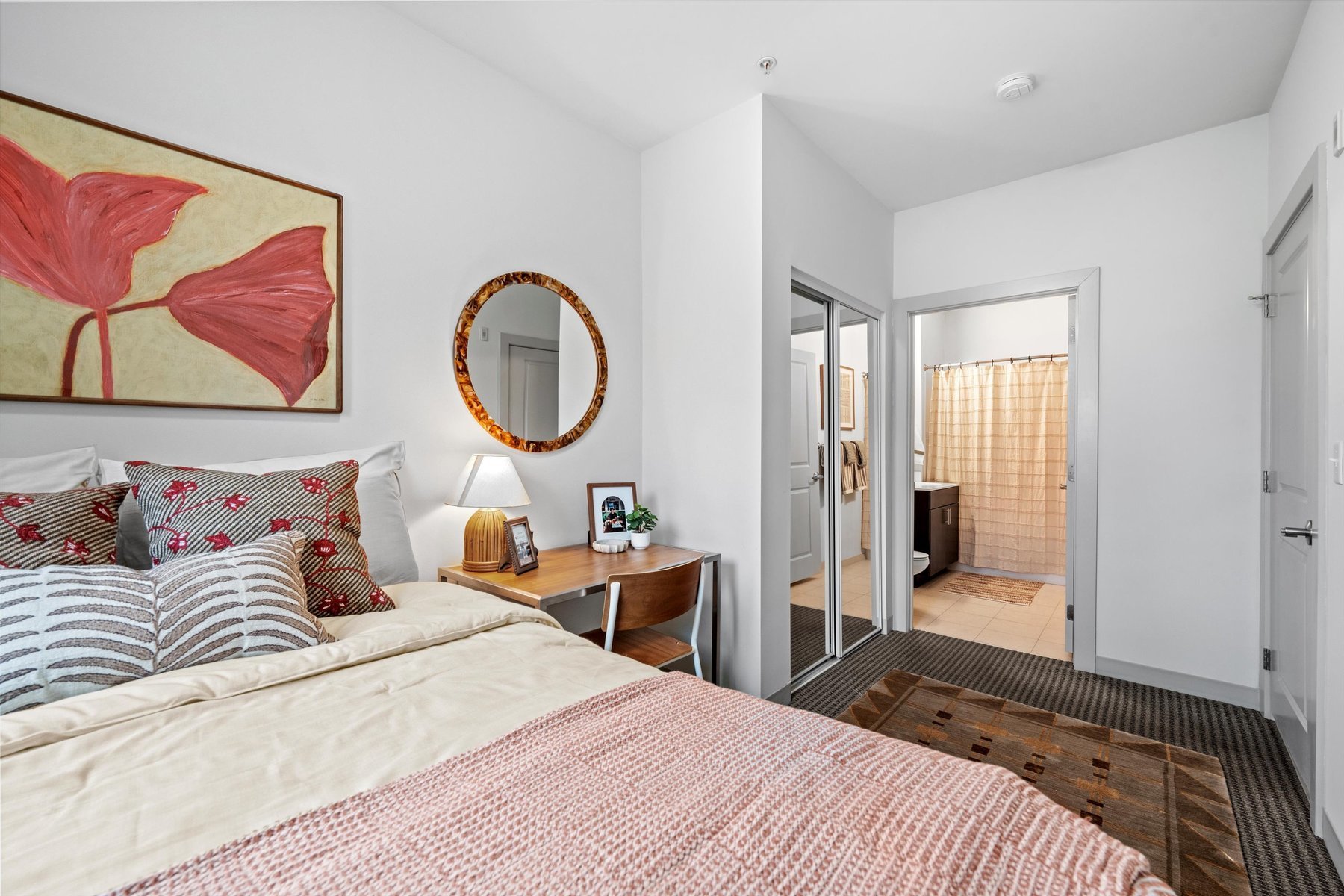 A cozy bedroom at Rambler College Park South features a neatly made bed, patterned pillows, a wooden desk with a lamp and framed photo, a round mirror, and a large painting above the bed; a doorway leads to a bright bathroom.