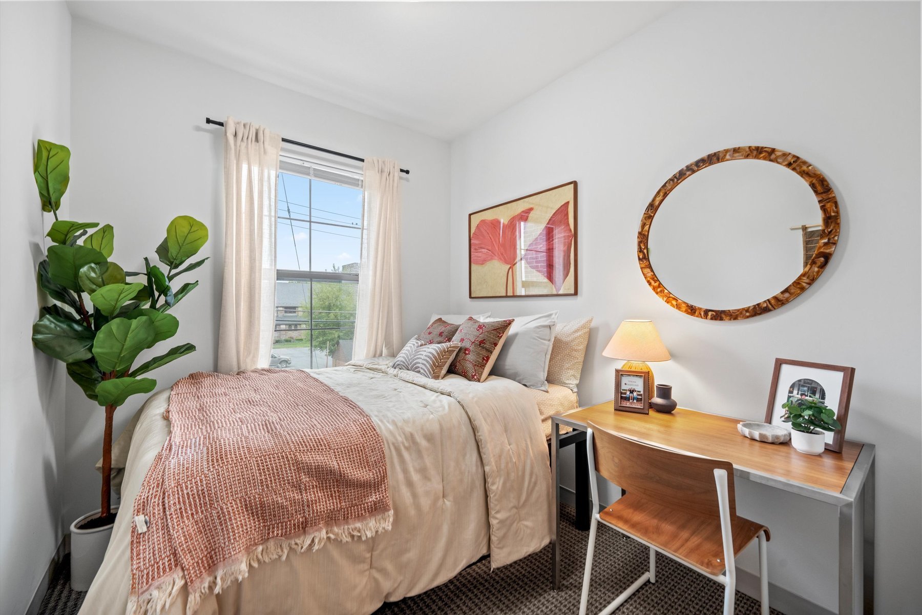 A cozy bedroom at Rambler College Park South features a neatly made bed, a large green plant, a window with sheer curtains, a desk with a lamp and photo frame, a wooden chair, and wall art with red leaves and a round mirror.