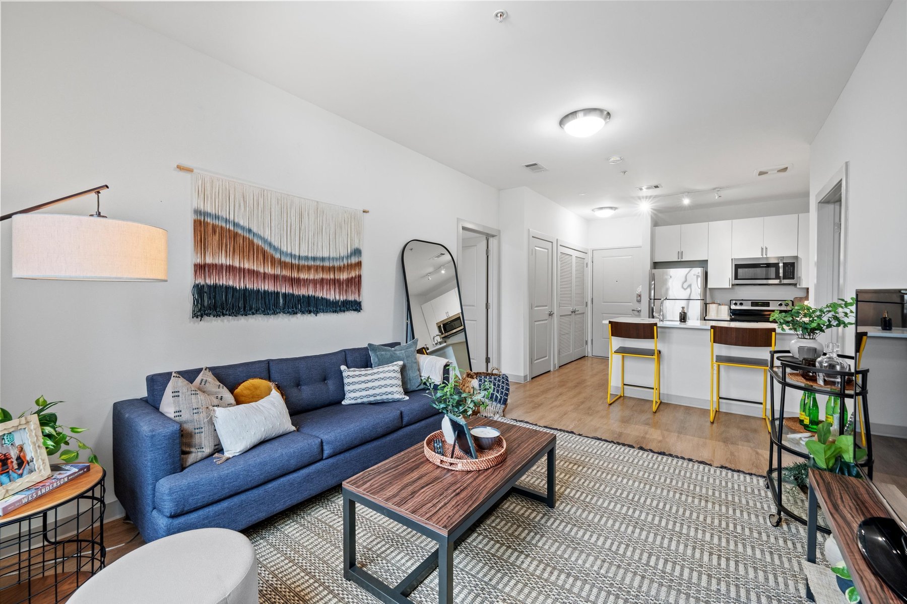 Modern open-concept living room and kitchen at Rambler College Park South with a blue sofa, round mirror, and cozy decor. The kitchen features white cabinets, stainless steel appliances, and two yellow barstools at the island.