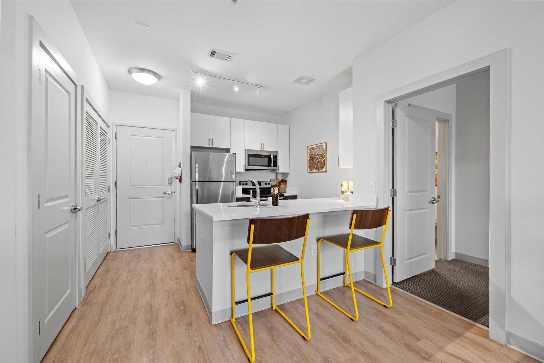 Modern kitchen at Rambler College Park South with white cabinets, stainless steel appliances, an island with yellow bar stools, light wood flooring, and open doors to adjacent rooms. Bright, clean, and minimalistic design.