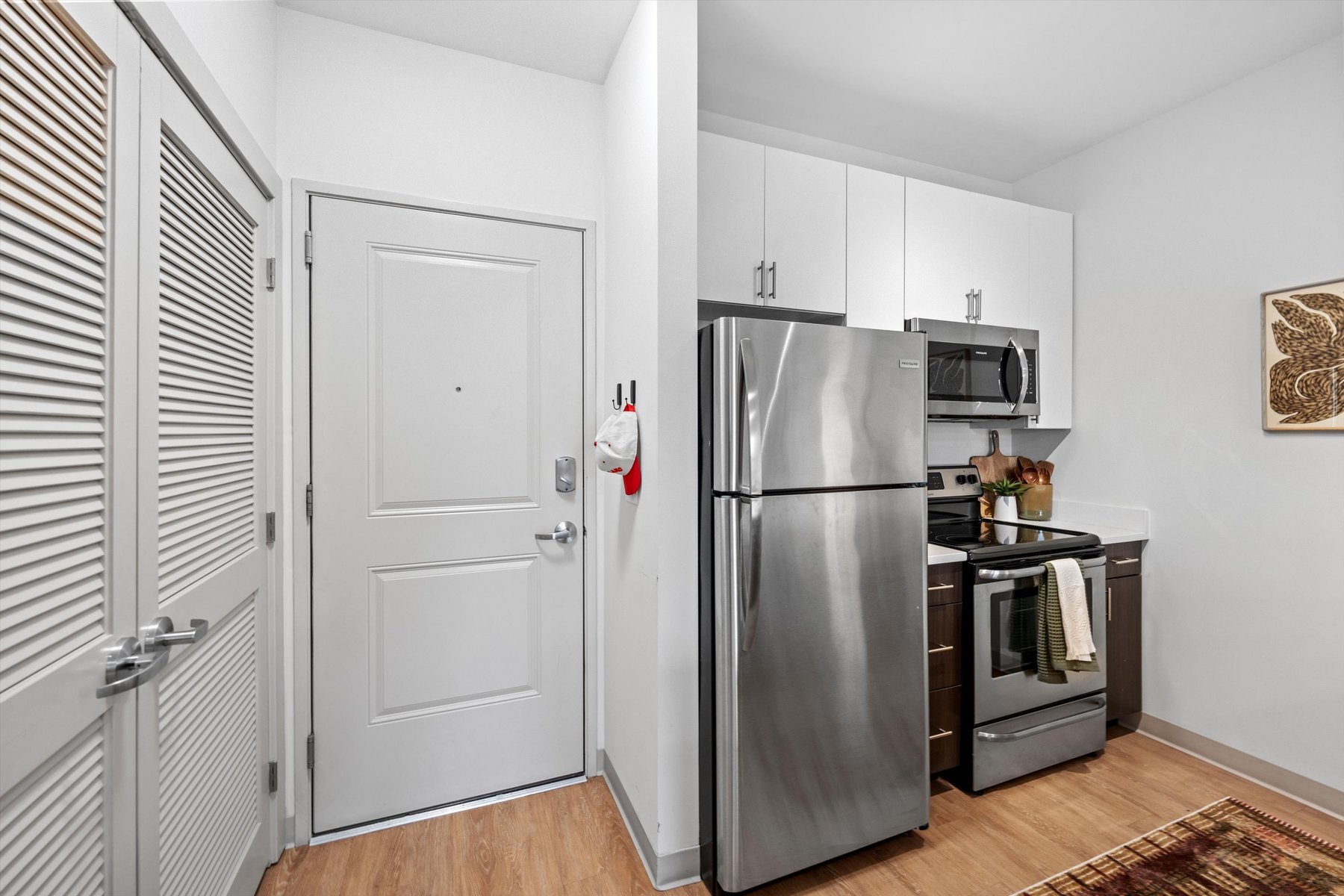 Modern kitchen at Rambler College Park South with stainless steel refrigerator, oven, and microwave, white cabinets, wooden floor, and a closed white entry door with hooks holding hats next to the kitchen area.