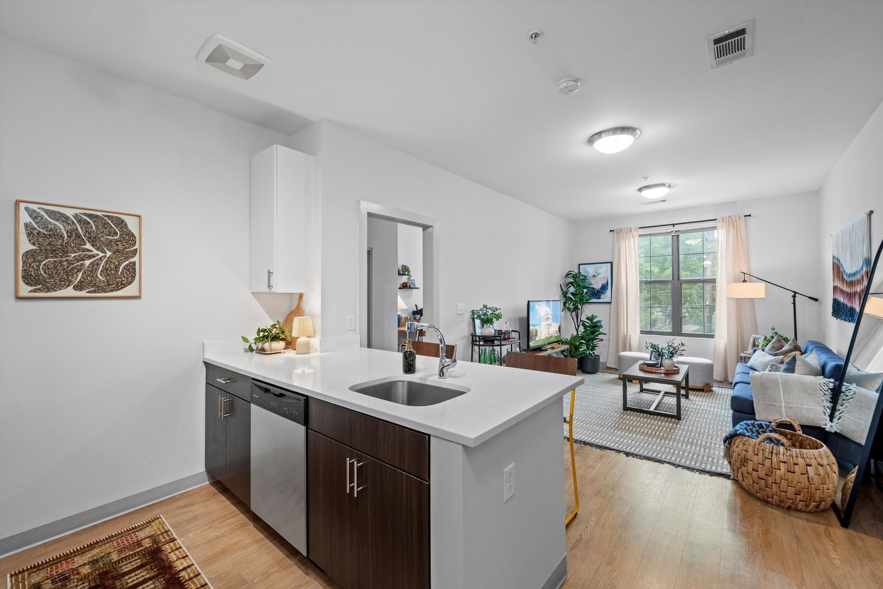 Modern open-concept apartment at Rambler College Park South with a white kitchen island and stainless steel sink, leading into a bright living room featuring a blue sofa, plants, large window, and plenty of natural light.
