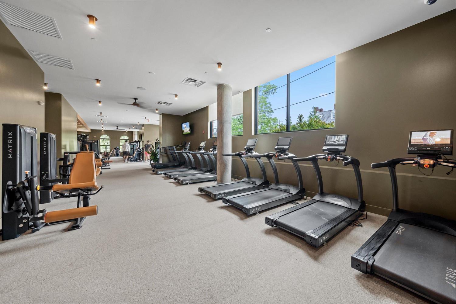 A modern gym at Rambler College Park South features treadmills lined up by windows, strength training machines on the left, TVs mounted on the walls, and natural light streaming in from large windows.