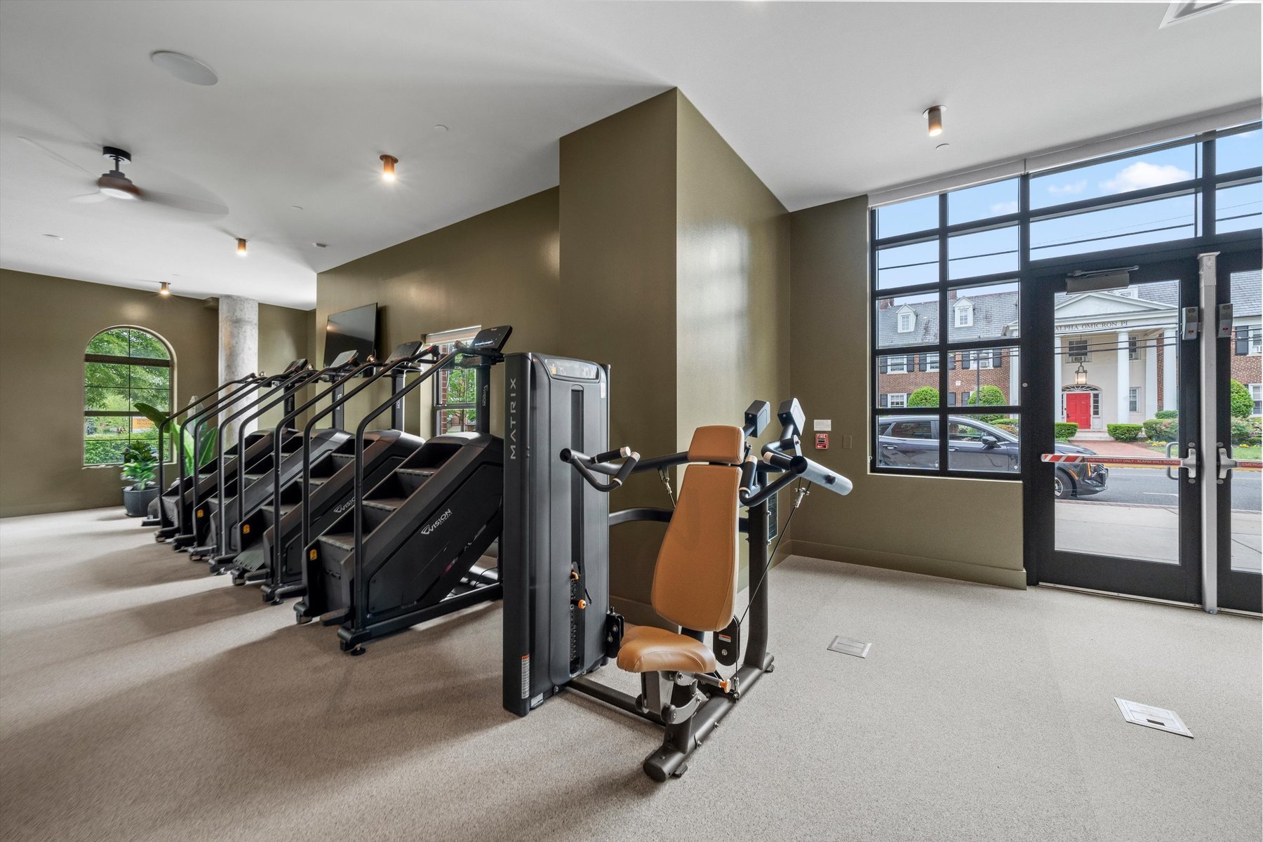A modern gym at Rambler College Park South with brown walls features several treadmills and a seated exercise machine near large windows and a glass door overlooking buildings and parked cars outside.