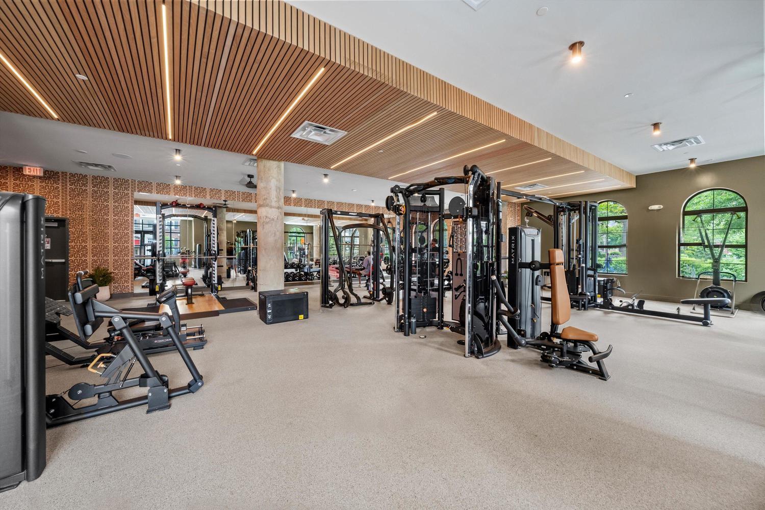 Spacious gym at Rambler College Park South with exercise machines, weight equipment, mirrors, and large windows letting in natural light. The room features modern decor and stylish wood-paneled ceiling accents.