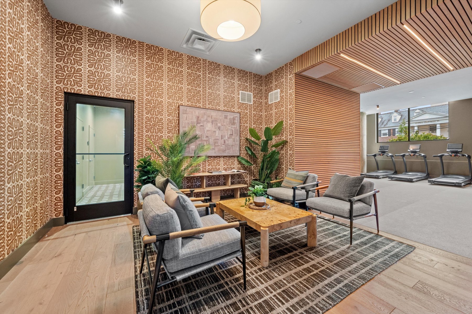 Modern lounge area with patterned walls, gray armchairs, a wooden coffee table, potted plants, and a rug at Rambler College Park South; adjacent to a fitness room with treadmills visible through an open wooden slat partition.