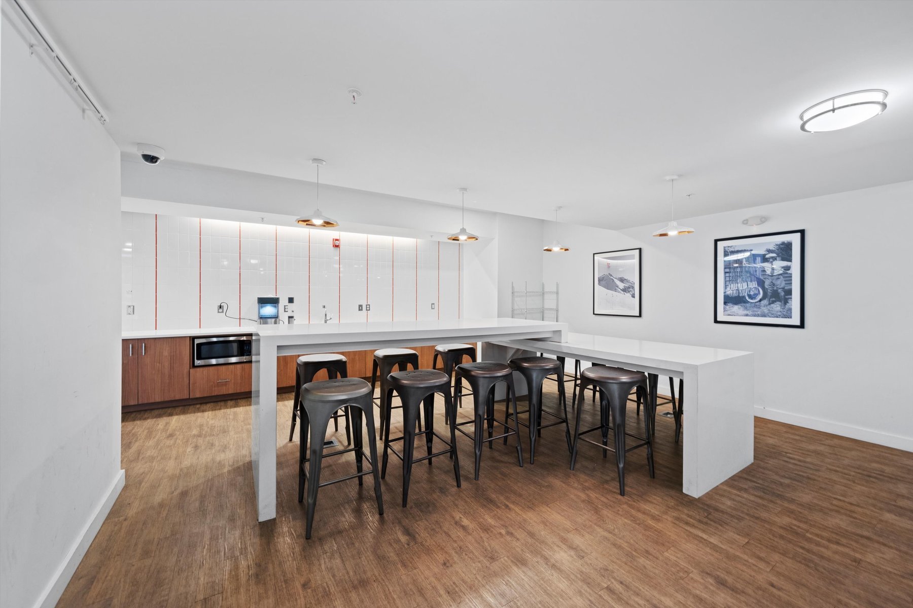 Modern break room at Rambler College Park South with white walls, a long white table, black metal stools, wooden floors, and a kitchenette with microwave. Pendant lights illuminate the space as two large framed photographs decorate the wall.