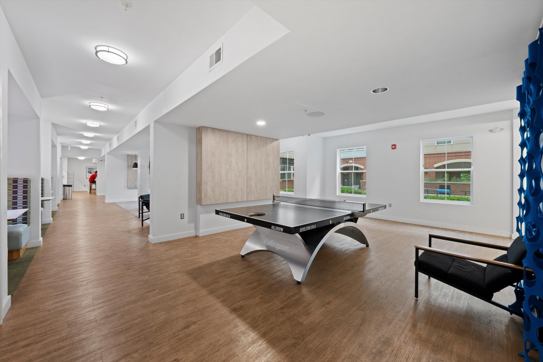 A modern recreation room at Rambler College Park South features wood flooring, a black ping pong table in the center, large windows streaming natural light, a black chair, and a hallway visible to the left.