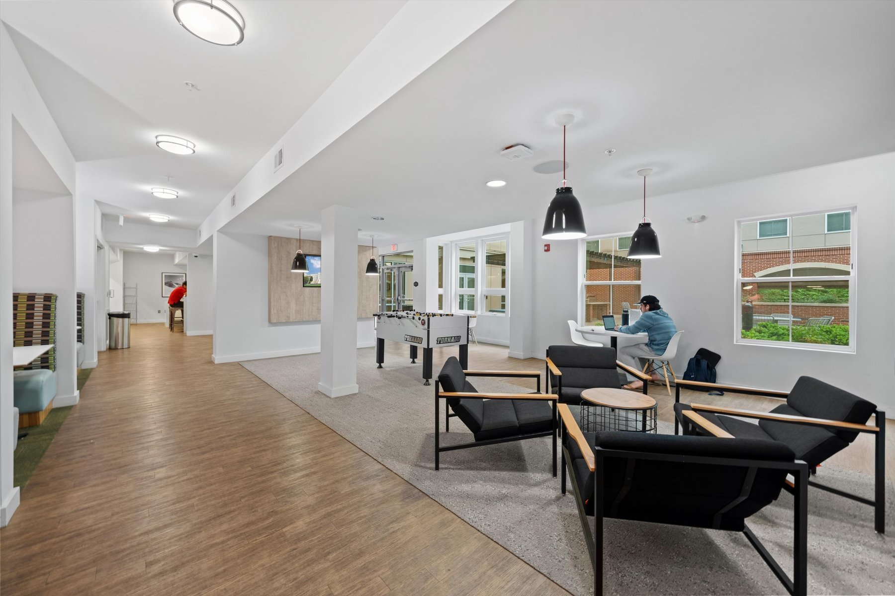 Modern common area at Rambler College Park South with black armchairs, tables, pendant lights, and large windows; one person working on a laptop at a table, another walking near the foosball table in the background.