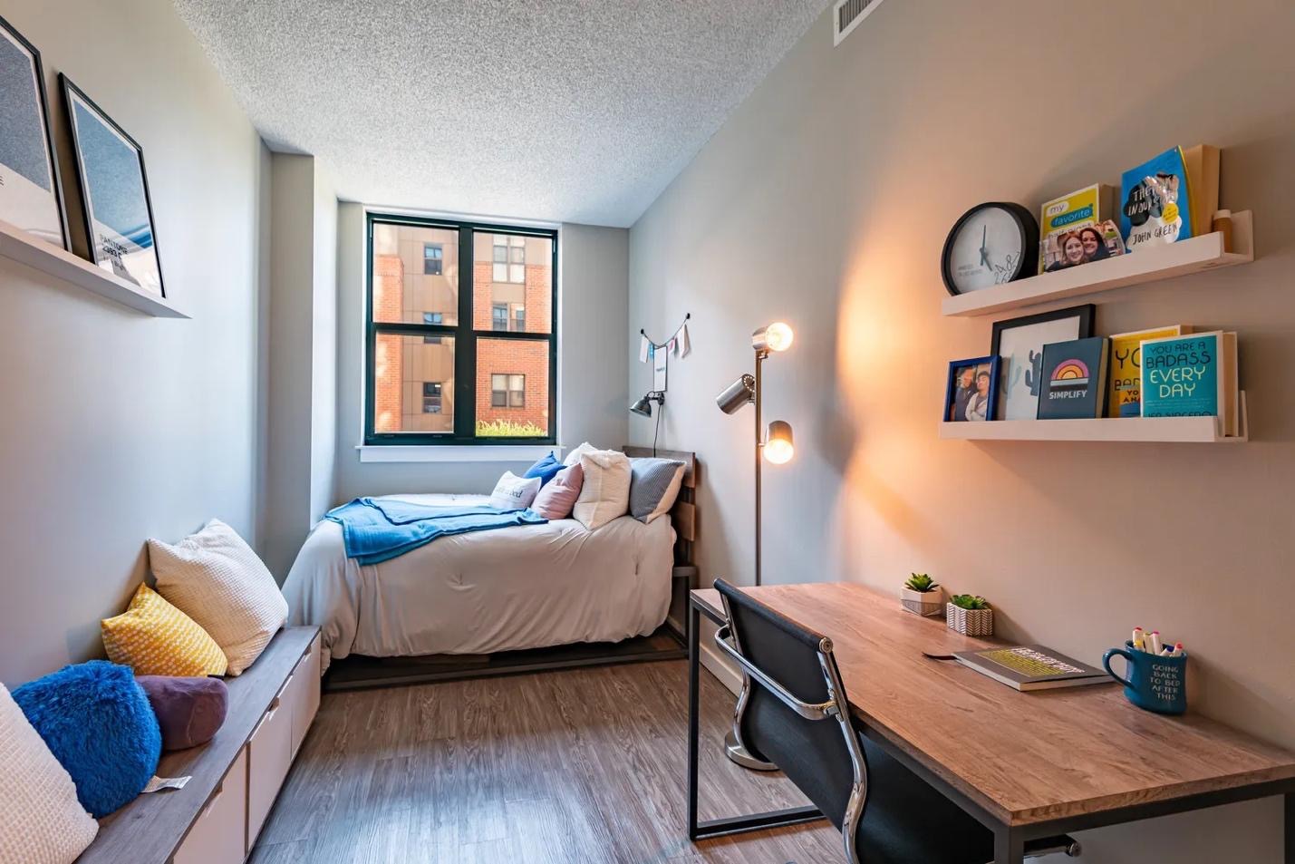 A cozy, modern dorm room like those in the best apartments near UMD—featuring a neatly made bed by the window, wall art, a bookshelf with photos, a comfy bench with pillows, and a wooden desk with a lamp, clock, and plant.