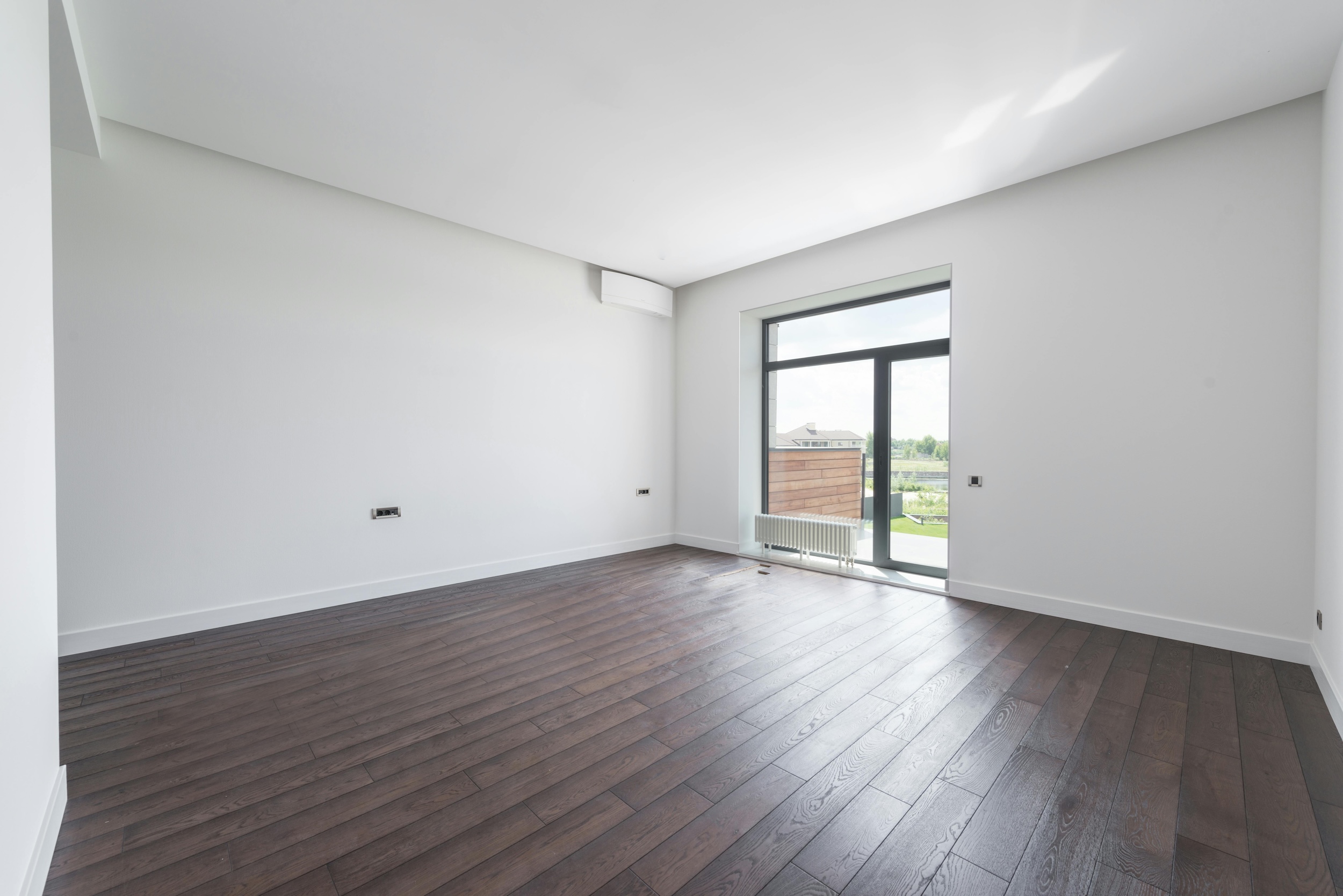 Empty room with white walls, dark wooden floor, a large window with a door leading outside, and sunlight streaming in.