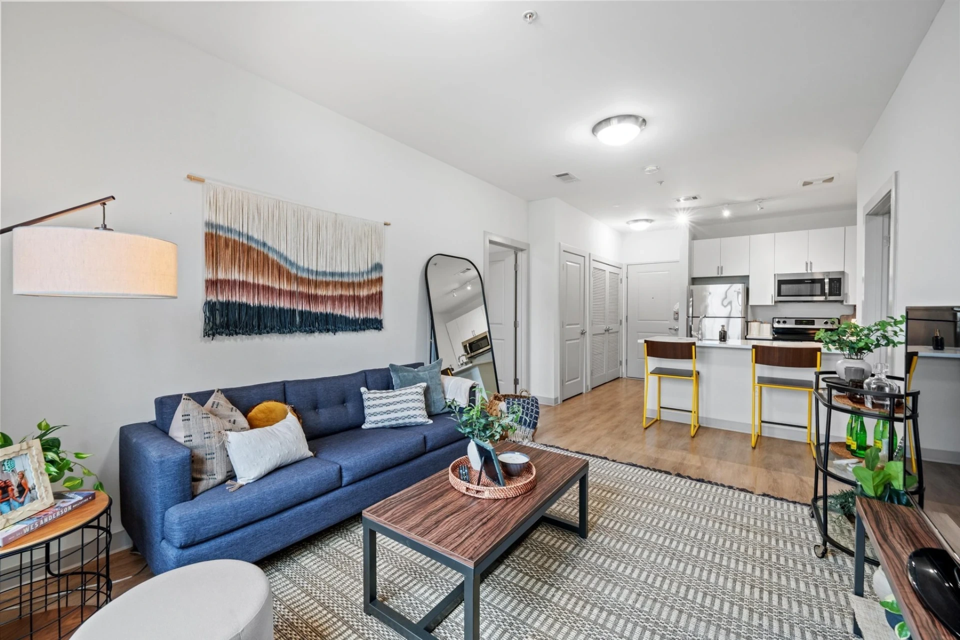 Modern open-plan living room and kitchen with a blue sofa, colorful wall hanging, wooden coffee table, tall mirror, potted plants, and a kitchen island with two yellow barstools under bright ceiling lights.