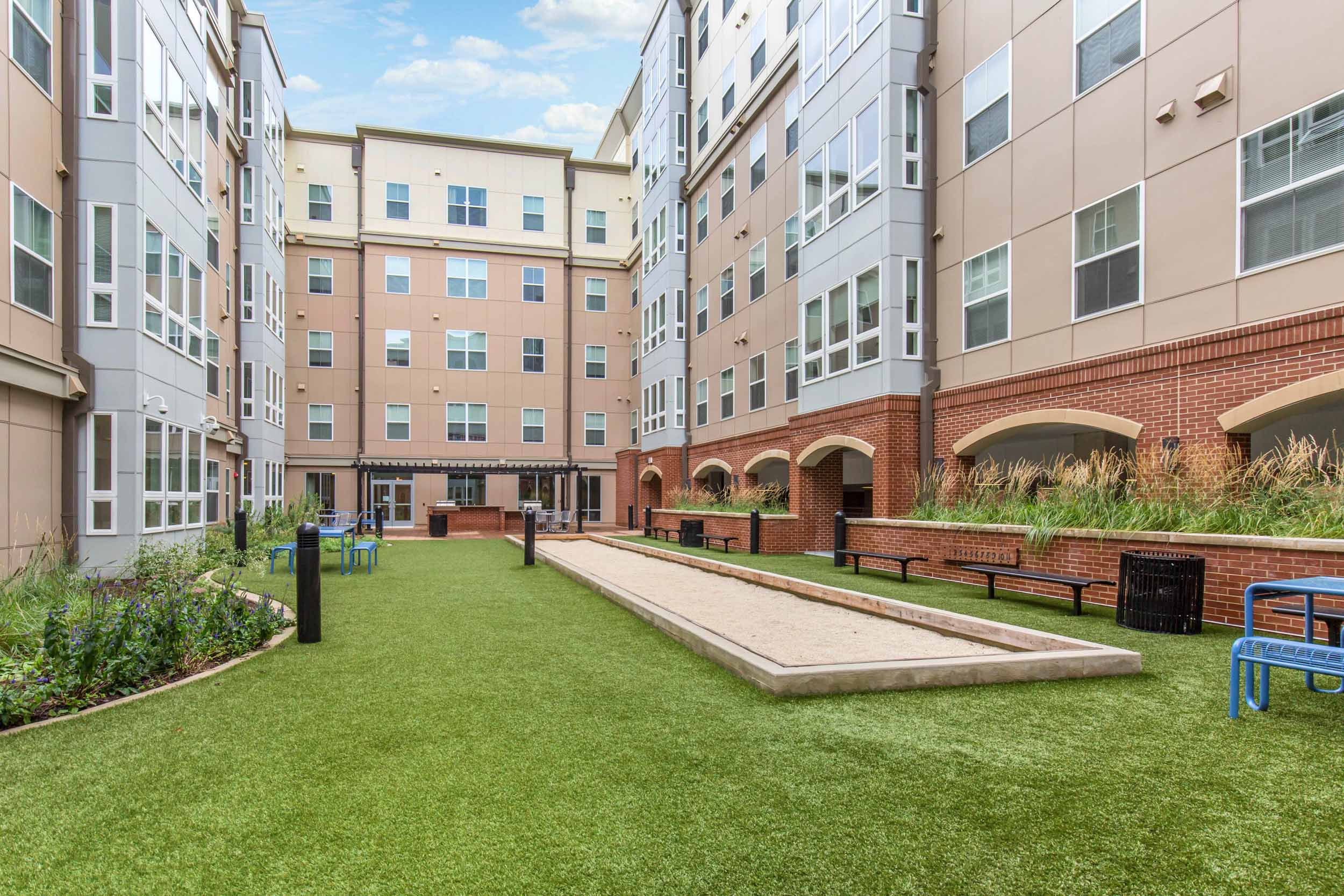 A courtyard with artificial grass, benches, a bocce court, landscaped plants, and tall apartment buildings surrounding the area under a partly cloudy sky.