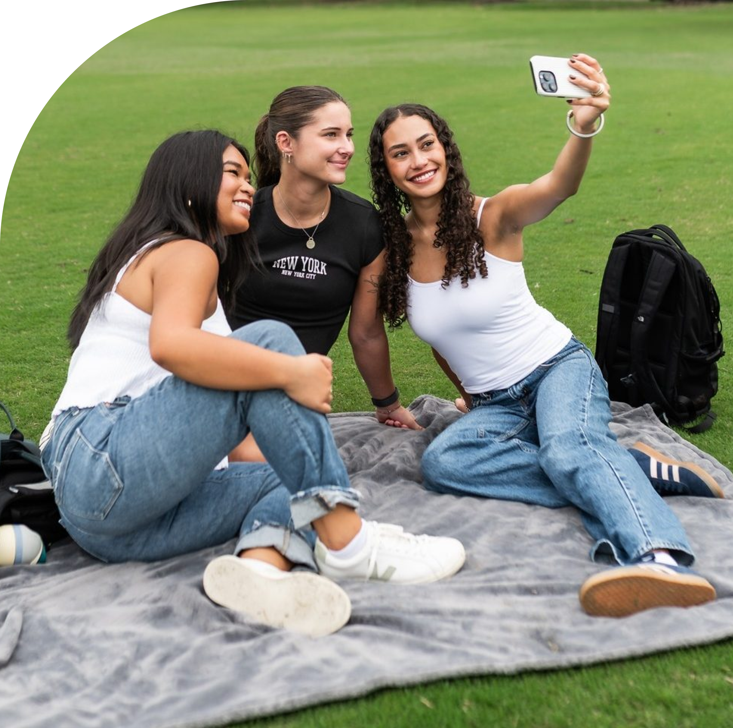 Three young women sit on a blanket in a grassy park, posing for a selfie. They are smiling, wearing casual clothes, and have backpacks nearby.