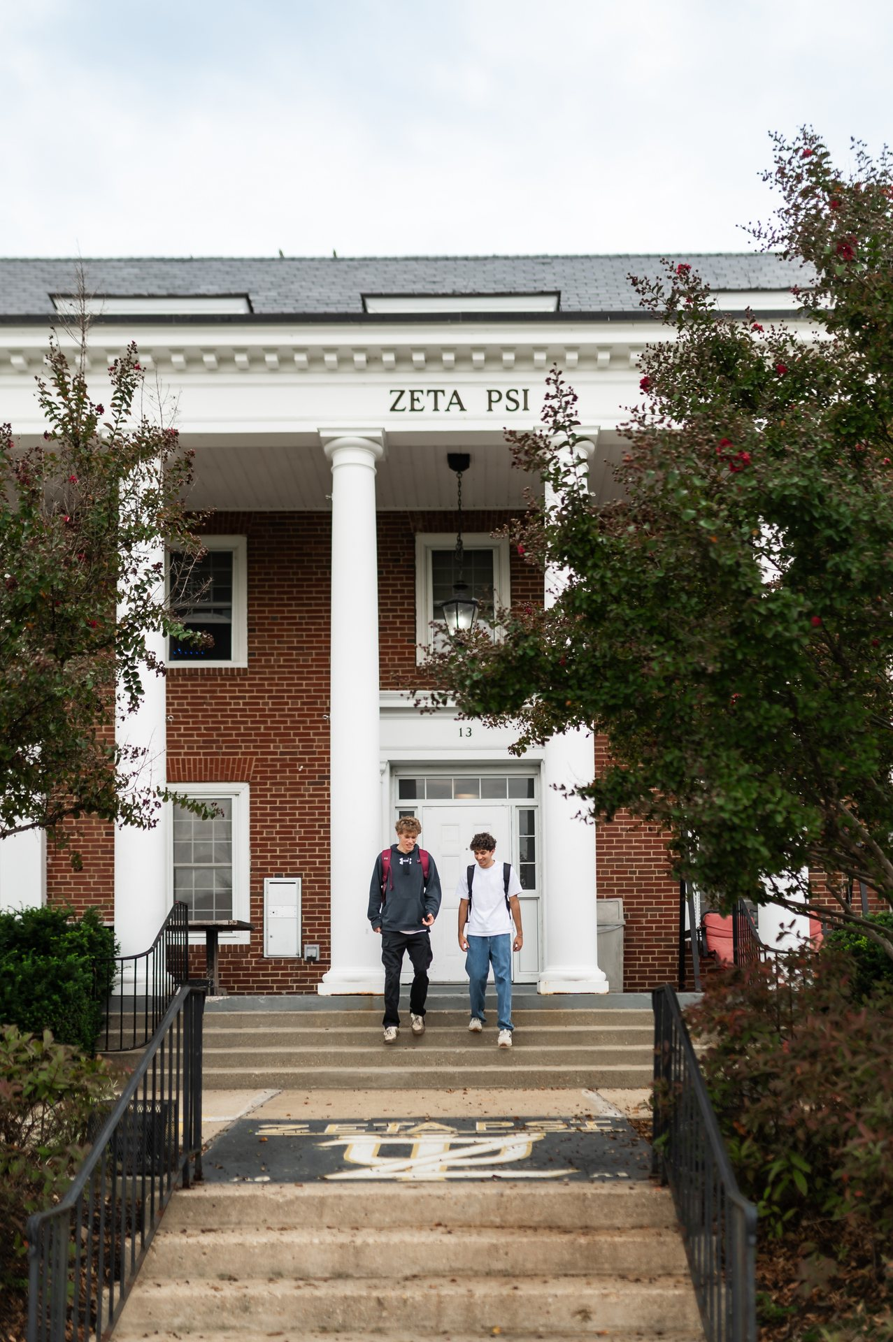 Two students walk down the steps outside a brick building with white columns labeled Zeta Psi. Trees frame the entrance, and a Greek letter symbol is painted on the walkway in front of the building.