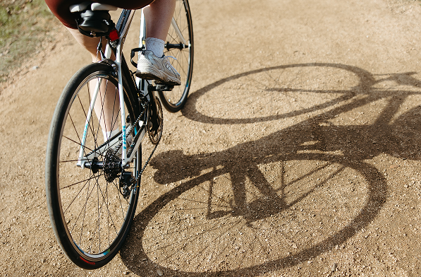 A person rides a bicycle on a dirt path near University of Maryland housing, casting a clear shadow on the ground. Only their legs and part of the bicycle are visible.