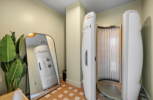 A standing tanning booth with open doors is next to a large mirror and a potted plant in a room with pale green walls and a patterned floor.