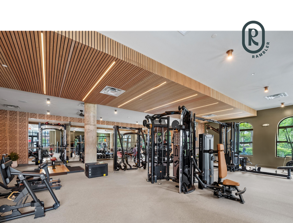 Spacious modern gym with various exercise machines, large mirrors, wood ceiling accents, and tall windows providing natural light—an inviting fitness space that exemplifies the amenities available in University of Maryland housing. Ramber logo in upper right corner.