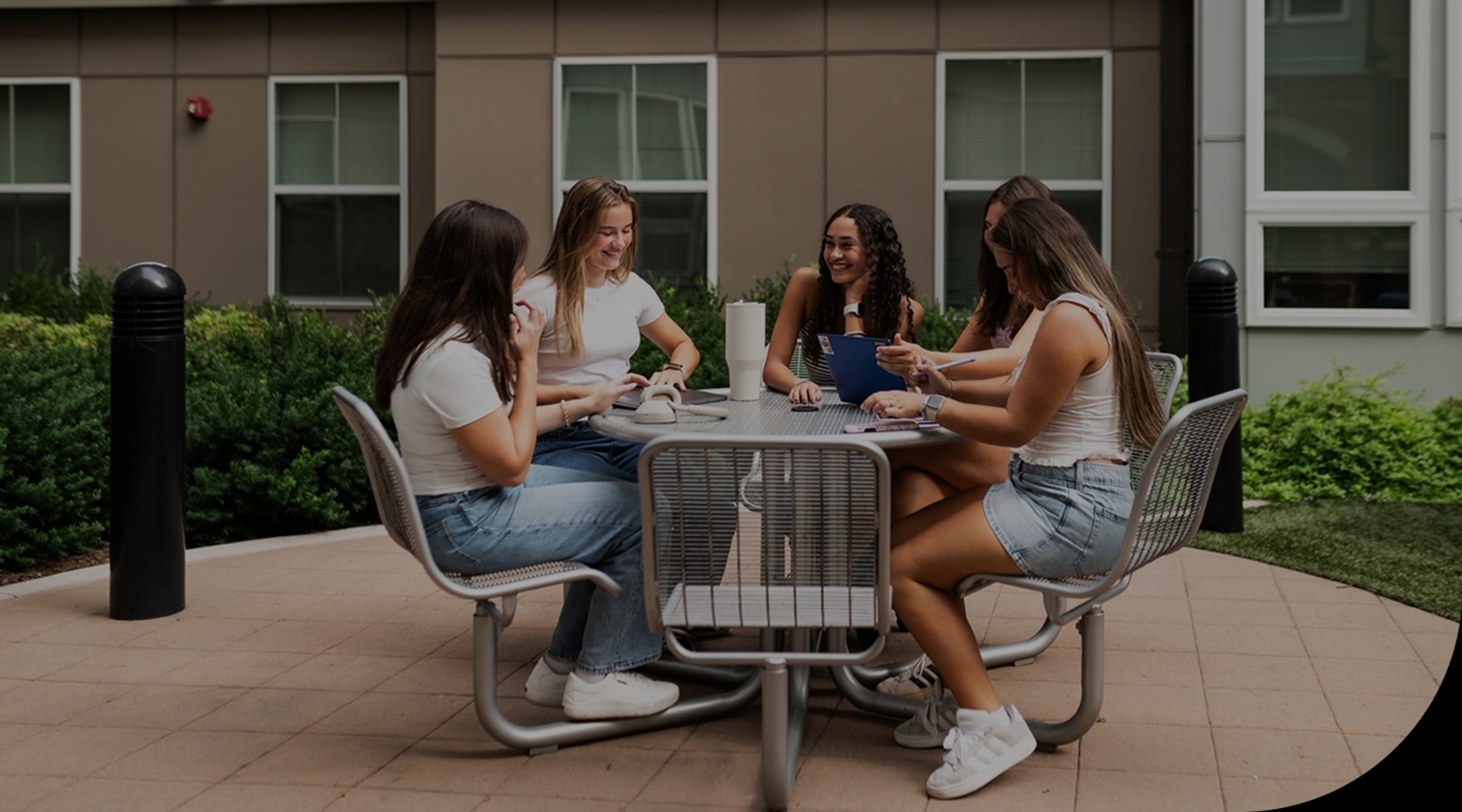 Five young women sit around an outdoor table, chatting and smiling. They are casually dressed in light clothing and sneakers. The background shows a building with large windows and greenery.