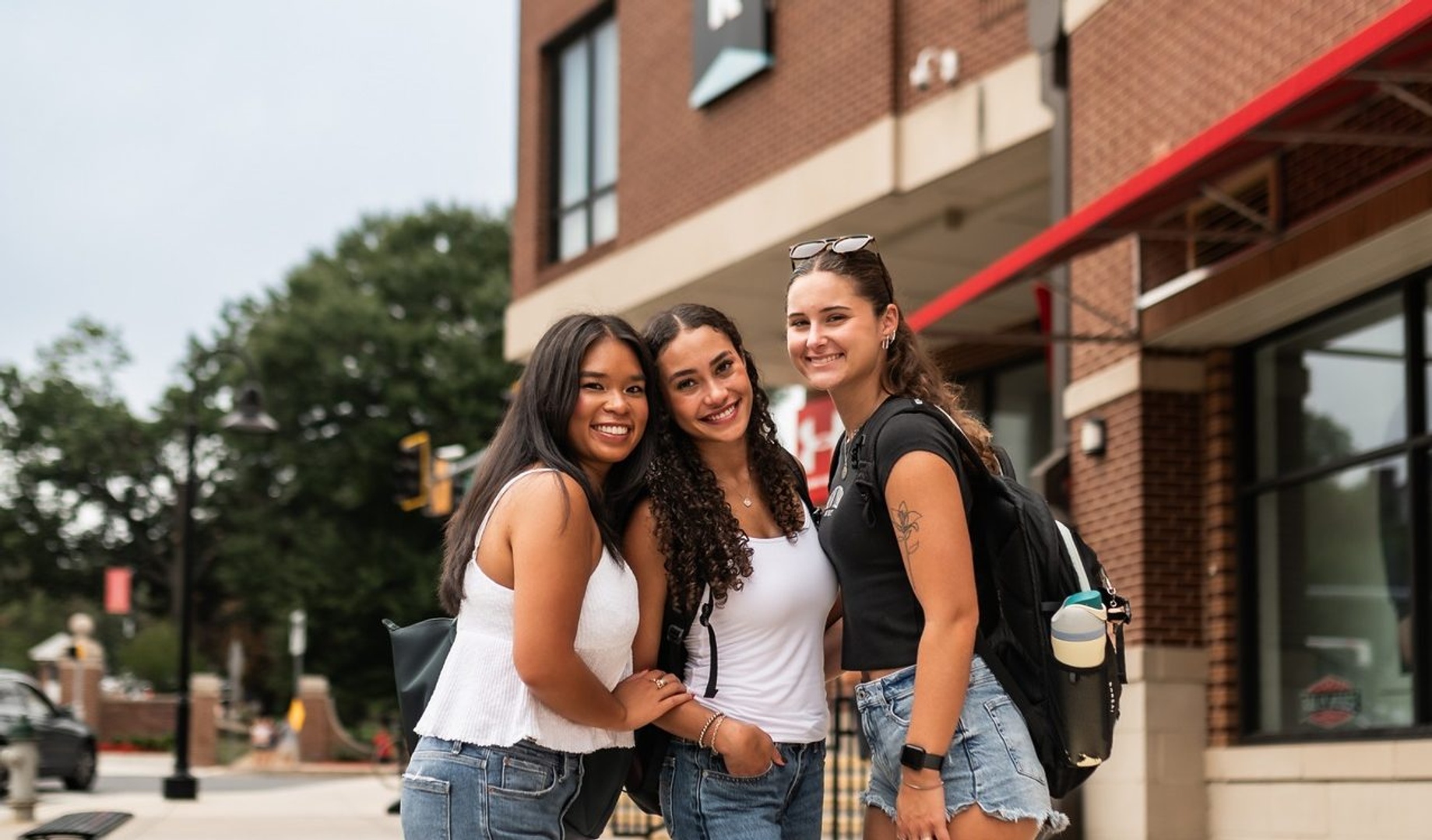 Three young women stand close together on a city sidewalk, smiling at the camera. They wear casual summer outfits with shorts and tank tops. The background shows a brick building and some greenery.