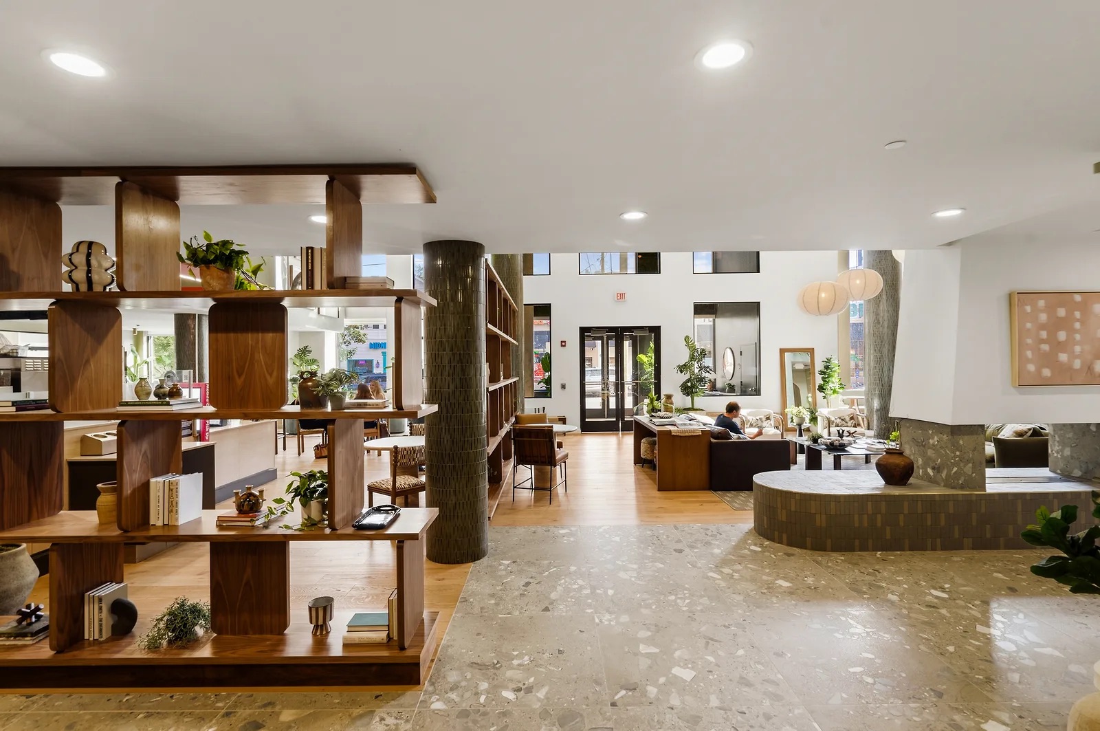 Modern open-concept lobby at Rambler College Park South with wooden shelves, lounge seating, large windows, plants, and artwork on the walls. People are relaxing and working in the bright, inviting space.