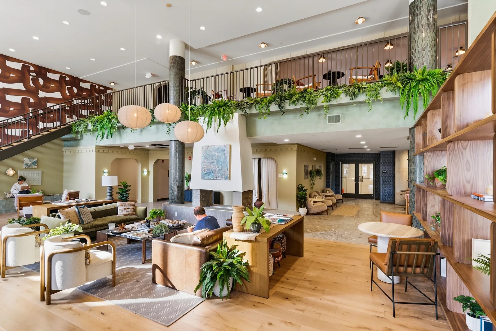 A modern, airy hotel lobby at Rambler College Park South features lush plants, wooden furniture, a mezzanine seating area, and large hanging lights. People are sitting and reading or conversing in this bright, welcoming space.