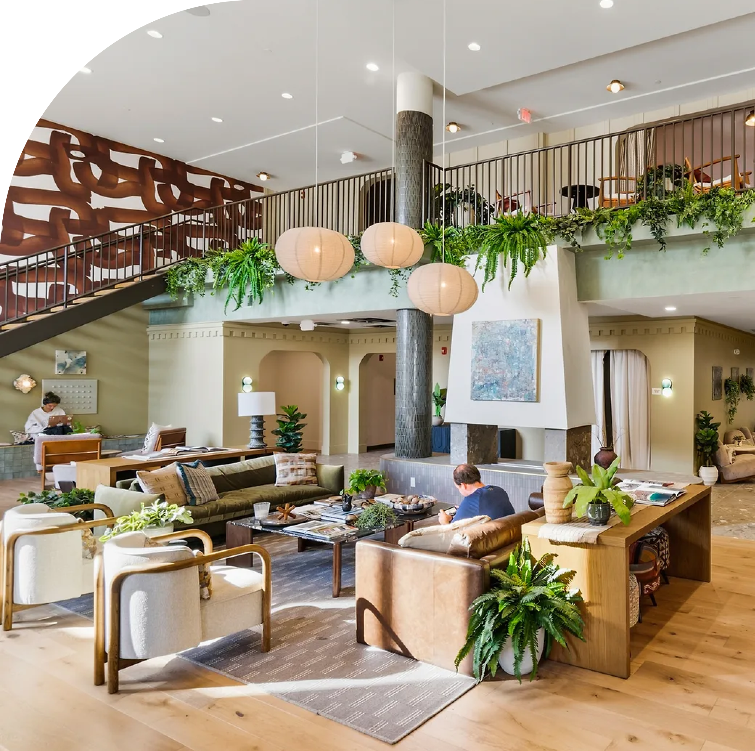 Modern, spacious hotel lobby with stylish furniture, plants, hanging lights, and an upper balcony—reminiscent of upscale University of Maryland housing. People are sitting and working in a bright, welcoming atmosphere.