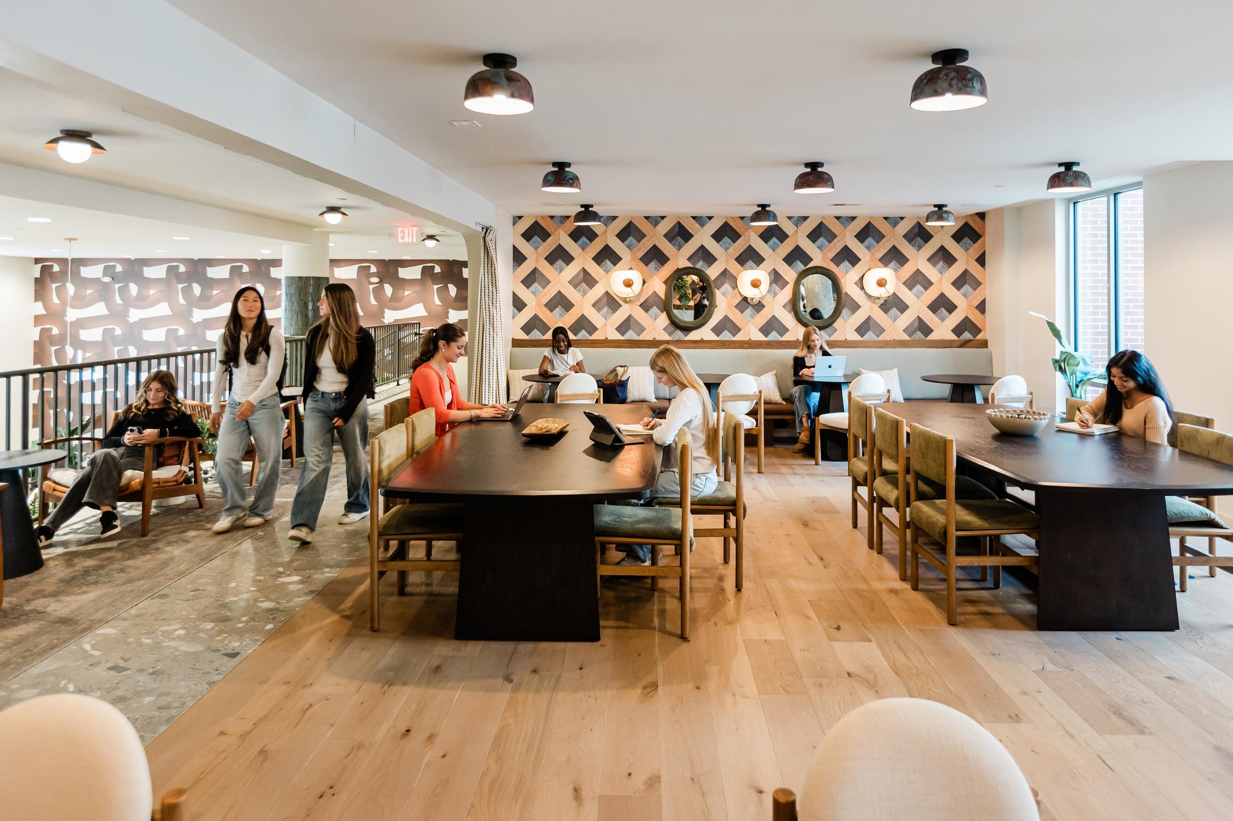 People study and read at dark wood tables in a modern, well-lit study area with geometric wall decor in Rambler College Park South. Some individuals walk or sit in an adjacent lounge area, while large windows let in ample natural light.
