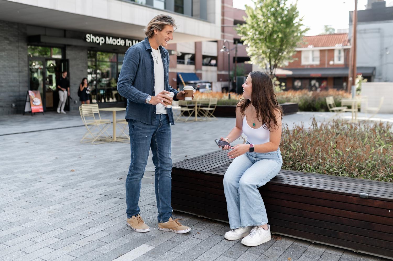 A man holding coffee cups smiles and chats about renter's insurance with a seated woman in an outdoor plaza filled with tables and plants. Both are casually dressed and appear to be enjoying their conversation.