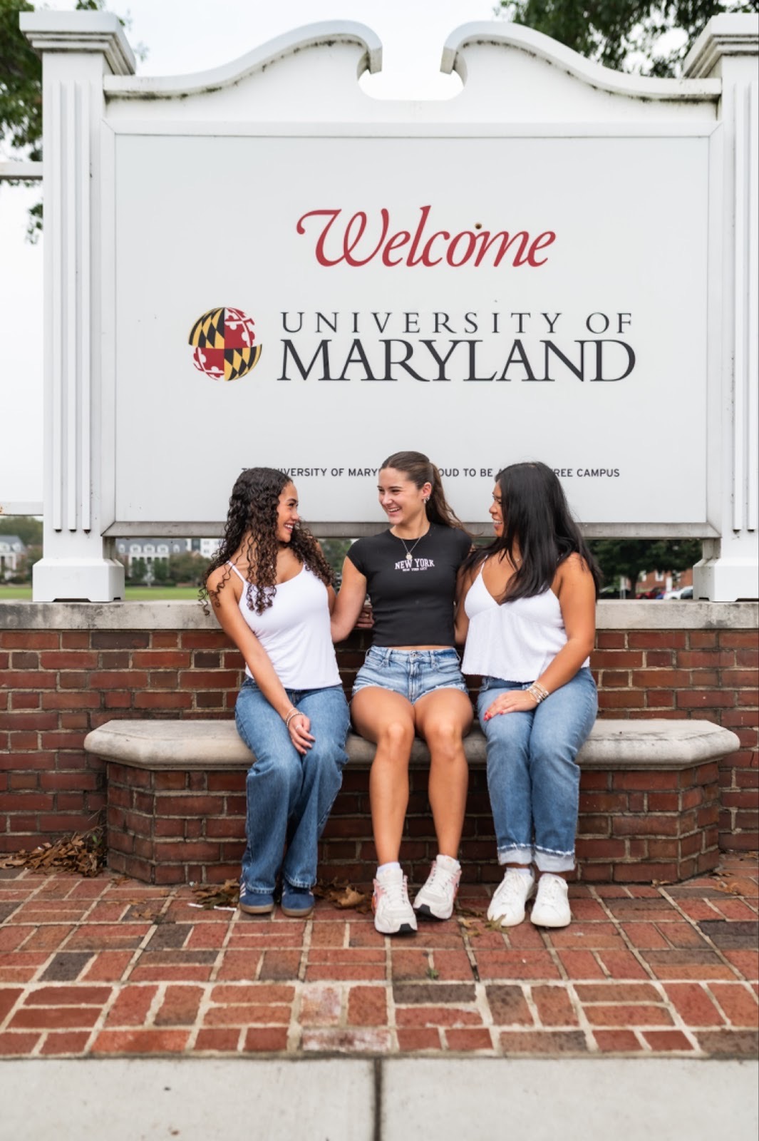 Three women sit on a curved bench in front of a Welcome University of Maryland sign, smiling and talking together on a brick-paved walkway, perhaps sharing tips about finding a roommate.