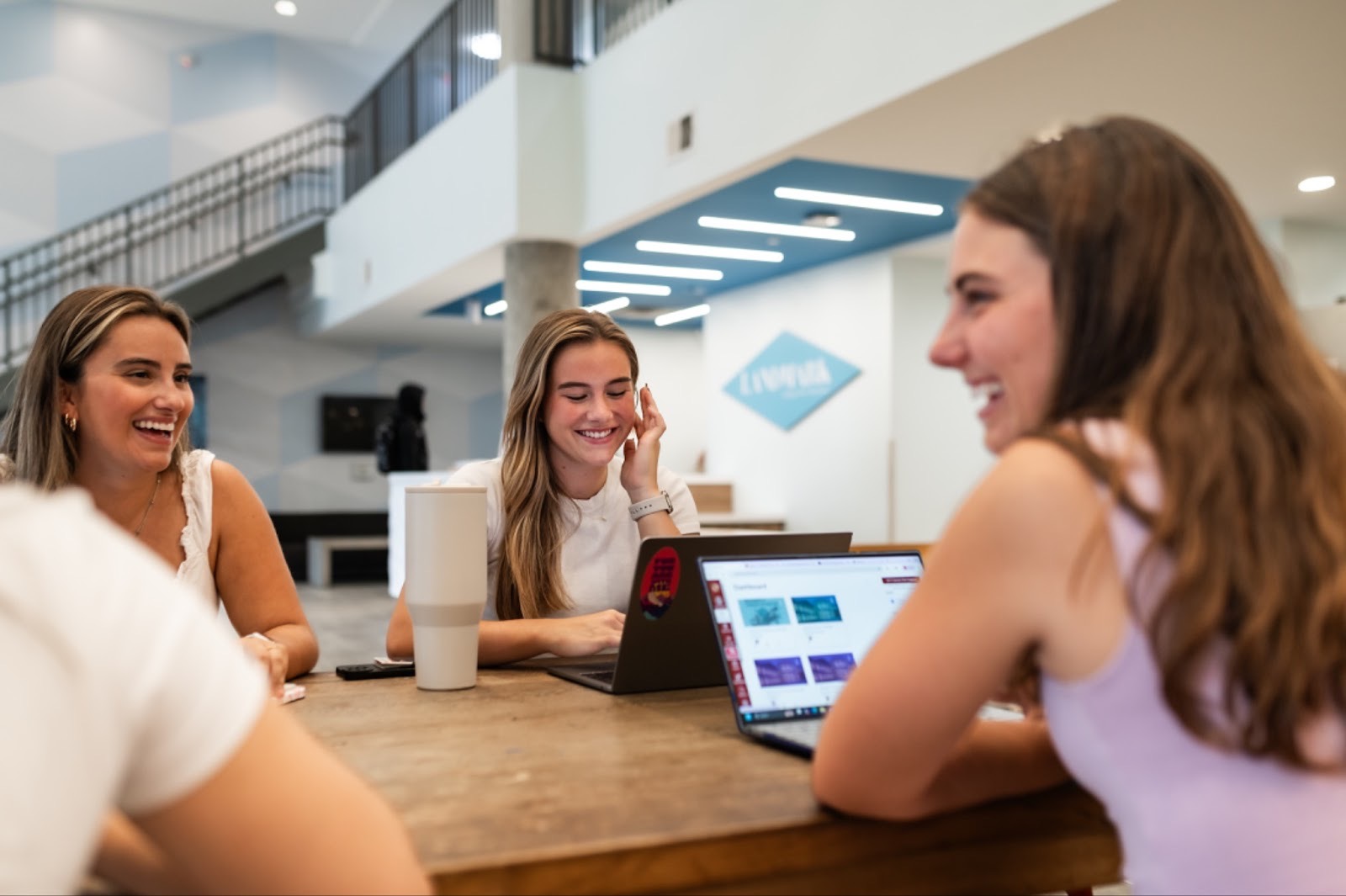 Three young women sit at a wooden table inside a modern building, smiling and talking, capturing the lively atmosphere in the best student apartments near UMD. Stairs and geometric wall patterns decorate the background.