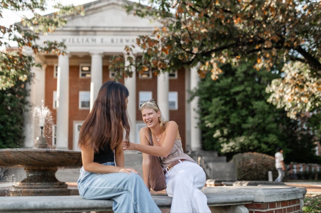 Two young women sit and laugh together on a stone bench in front of the William E. Kirwan building, a guarantor of campus tradition. A fountain and leafy trees frame the lively, sunny scene.