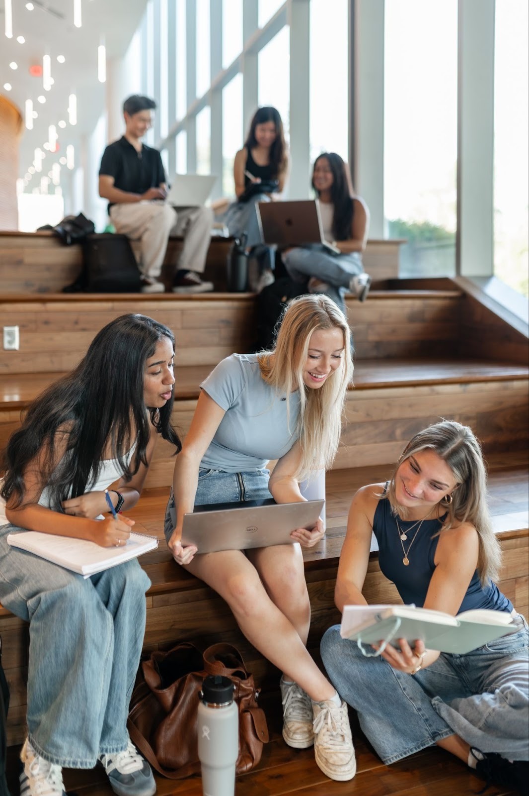 A group of students sits on wooden stairs, studying together. Three women in the foreground read a book and use a laptop, smiling. Others sit behind them, engaged in conversation and working on laptops near large windows.