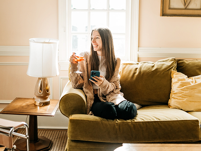 A woman sits on a green couch in her UMD Off-Campus Housing, smiling while holding a drink and a smartphone. Sunlight streams through the window behind her, brightening the cozy living room.